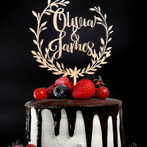 May include: A white cake with chocolate frosting and a wooden cake topper that says "Olivia & James" in cursive. The cake is decorated with fresh strawberries, blueberries, and raspberries.