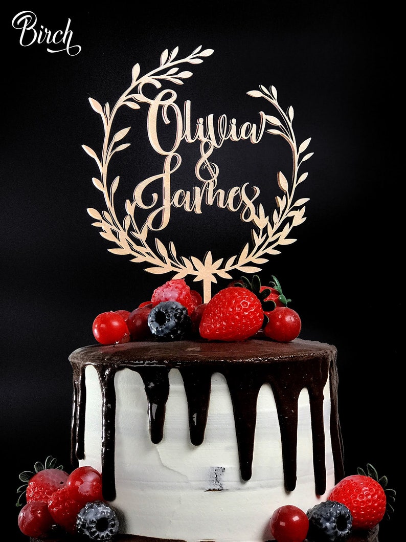 May include: A white cake with chocolate frosting and a wooden cake topper that says "Olivia & James" in cursive. The cake is decorated with fresh strawberries, blueberries, and raspberries.