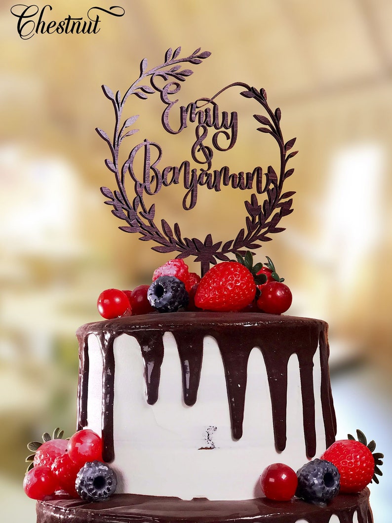 May include: A two-tiered wedding cake decorated with chocolate frosting and fresh berries. The cake is topped with a wooden cake topper that says "Emily & Benjamin" in a cursive font.