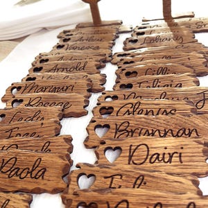Op de afbeelding: Houten plaatskaarten met hartvormige uitsnijdingen en gegraveerde namen. De namen omvatten Arnold, Vanessa, Lohany, Talia, Brisnman, Dairi en Nathalie.
