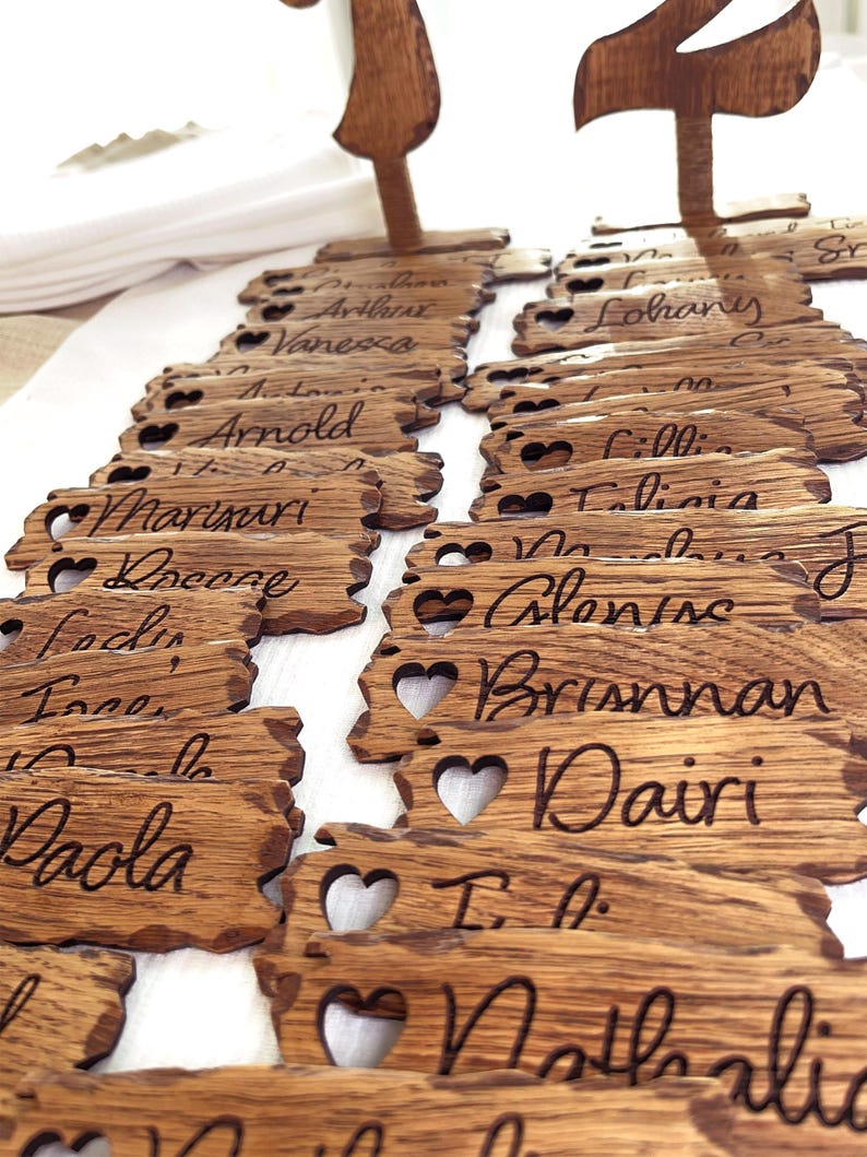 Op de afbeelding: Houten plaatskaarten met hartvormige uitsnijdingen en gegraveerde namen. De namen omvatten Arnold, Vanessa, Lohany, Talia, Brisnman, Dairi en Nathalie.