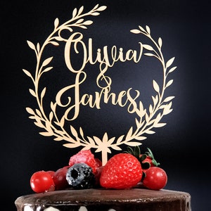 May include: A wooden cake topper with a leafy wreath design and the names "Olivia & James" in cursive script. The topper is placed on a chocolate cake decorated with fresh berries.