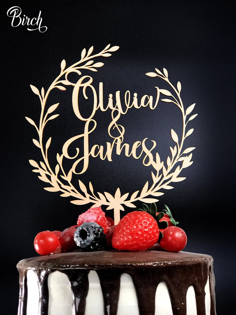 May include: A wooden cake topper with a leafy wreath design and the names "Olivia & James" in cursive script. The topper is placed on a chocolate cake decorated with fresh berries.