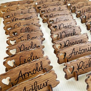 Op de afbeelding: Houten plaatskaarten met hartvormige uitsnijdingen en namen in cursief schrift. De namen omvatten Arthur, Arnold, Leslie, Paola, Lillian, Claire, Maverick, William, Nicholas,  en meer.