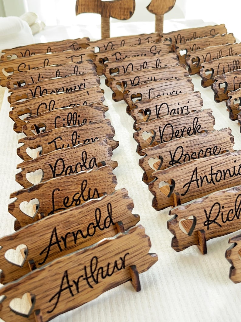 Op de afbeelding: Houten plaatskaarten met hartvormige uitsnijdingen en namen in cursief schrift. De namen omvatten Arthur, Arnold, Leslie, Paola, Lillian, Claire, Maverick, William, Nicholas,  en meer.