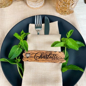 Op de afbeelding: Een zwart bord met een beige linnen servet, een houten plaatskaart met de naam "Charlotte" erop gegraveerd, en een vork en mes. De plaatskaart heeft een hartvormige uitsnijding. Het bord is omringd door takjes verse groene munt.