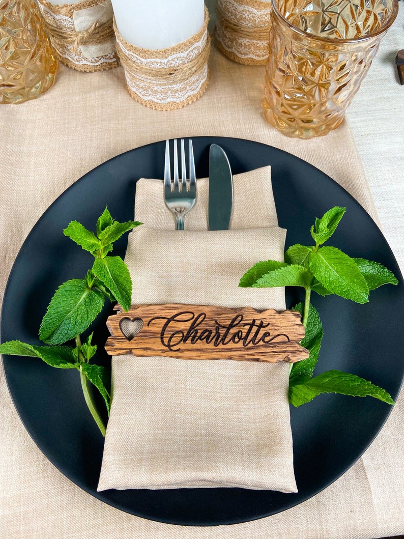 Op de afbeelding: Een zwart bord met een beige linnen servet, een houten plaatskaart met de naam "Charlotte" erop gegraveerd, en een vork en mes. De plaatskaart heeft een hartvormige uitsnijding. Het bord is omringd door takjes verse groene munt.