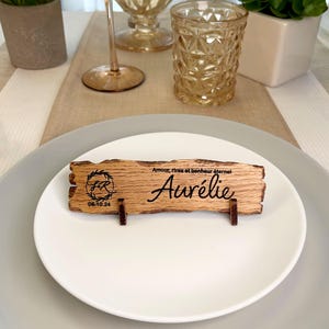 May include: A rustic wooden place card with the name "Aur&eacute;lie" engraved on it. The card also features a small wreath with the initials "F.R." and the date "08.10.24". The text "Amour, rires et bonheur &eacute;ternel" is also engraved on the card.
