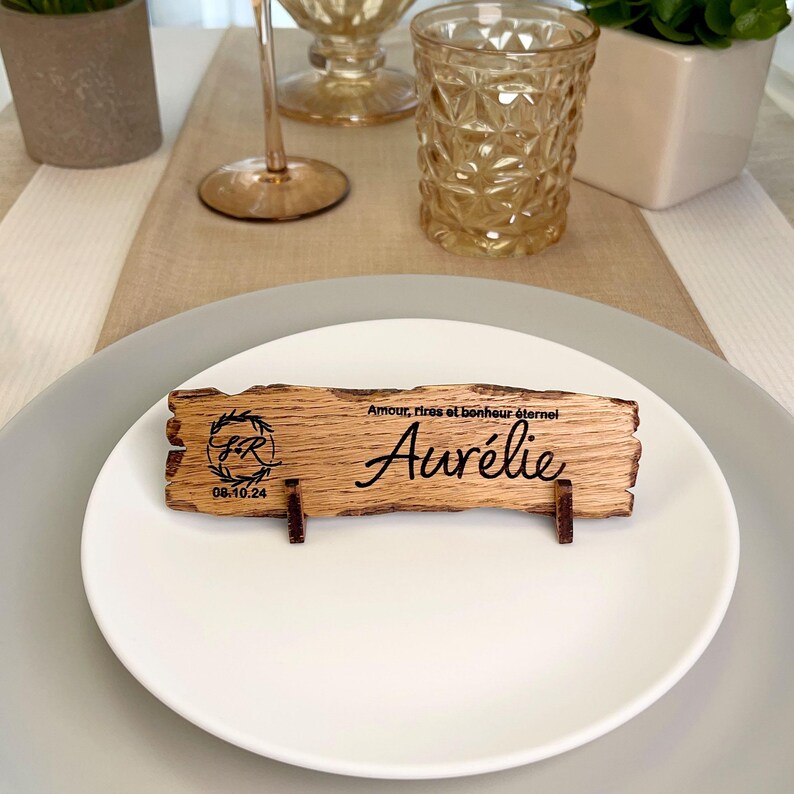 May include: A rustic wooden place card with the name "Aur&eacute;lie" engraved on it. The card also features a small wreath with the initials "F.R." and the date "08.10.24". The text "Amour, rires et bonheur &eacute;ternel" is also engraved on the card.