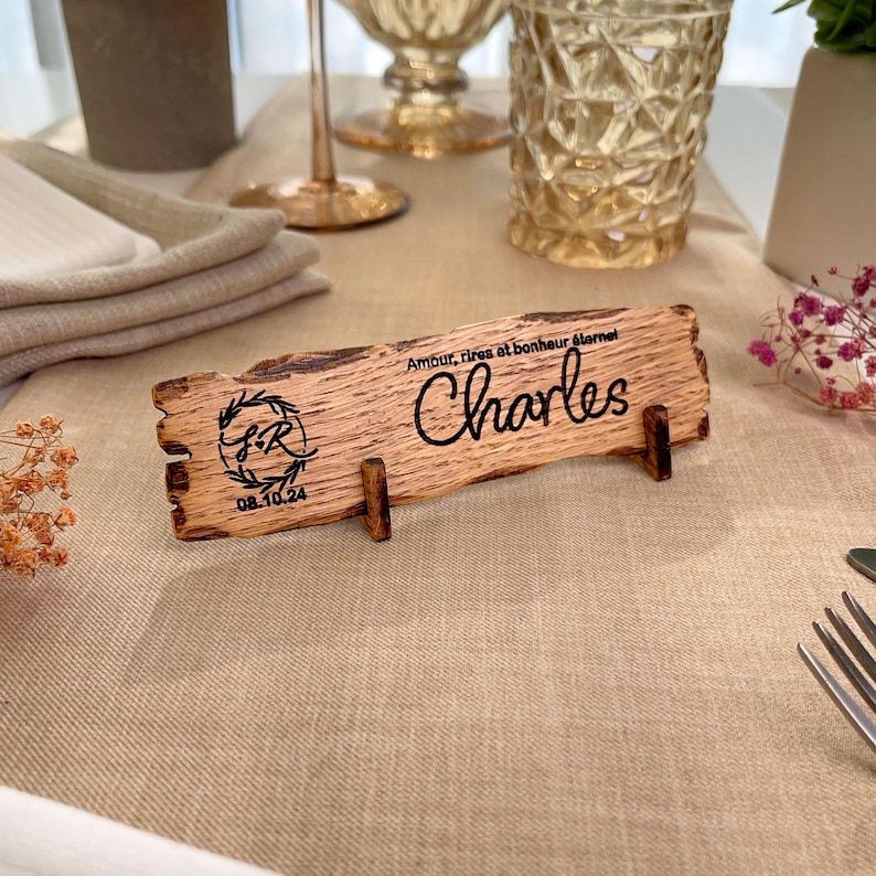 May include: A wooden table place card with the name "Charles" engraved on it. The card also features a floral design with the initials "S&R" and the date "08.10.24". The text "Amour, rires et bonheur &eacute;ternel" is also engraved on the card.