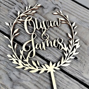 May include: A wooden cake topper with a leafy wreath design. The names "Olivia & James" are written in cursive inside the wreath.