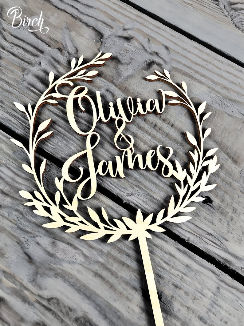 May include: A wooden cake topper with a leafy wreath design. The names "Olivia & James" are written in cursive inside the wreath.