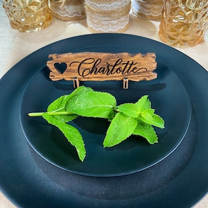 Op de afbeelding: Een houten plaatskaart met de naam "Charlotte" erop gegraveerd. De plaatskaart ligt op een zwart bord met een takje groene munt.
