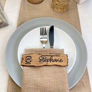 May include: A wooden place card with the name "Stephane" engraved on it. The card is decorated with a small circle containing the letters "FR" and the date "08.10.24". The card is placed on a linen napkin with a fork and knife.