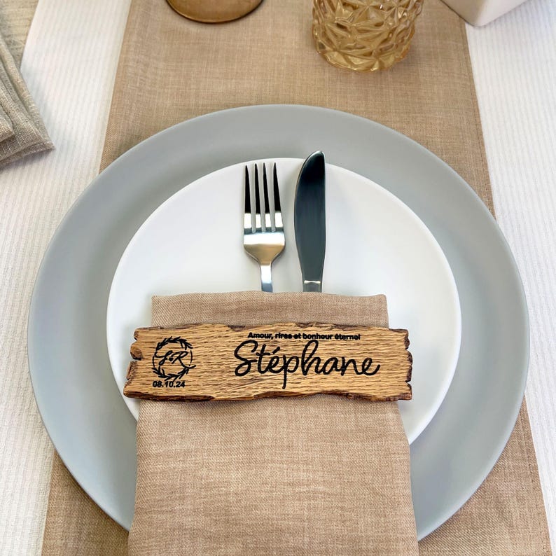 May include: A wooden place card with the name "Stephane" engraved on it. The card is decorated with a small circle containing the letters "FR" and the date "08.10.24". The card is placed on a linen napkin with a fork and knife.