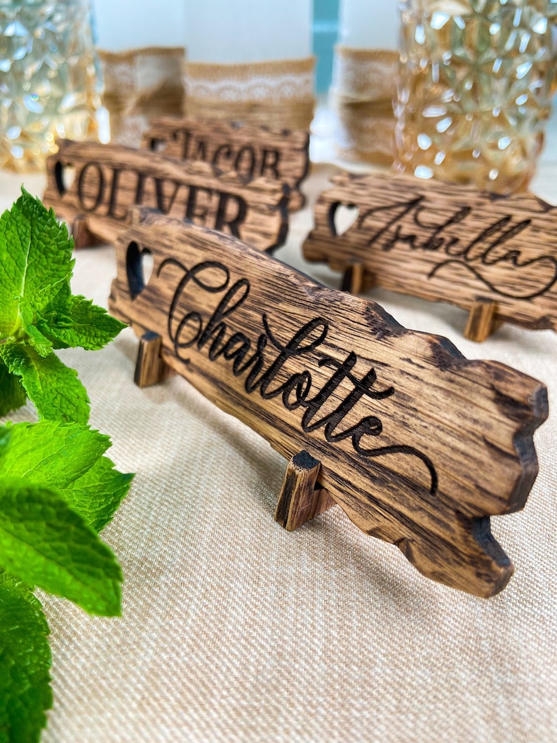 May include: Wooden table place cards with names "Jacob", "Oliver", "Isabella", and "Charlotte" written in cursive. The cards are on a beige linen tablecloth.