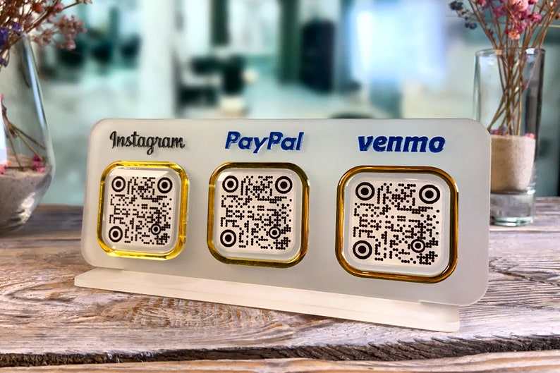 May include: White acrylic sign with three gold-rimmed QR codes. The sign has the words "Instagram", "PayPal", and "venmo" printed on it.