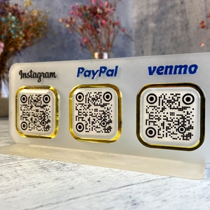 May include: A white acrylic sign with three gold-rimmed QR codes. The sign has the words "Instagram", "PayPal", and "Venmo" printed in blue.