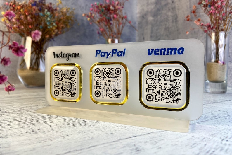 May include: A white acrylic sign with three gold-rimmed QR codes. The sign has the words "Instagram", "PayPal", and "Venmo" printed in blue.