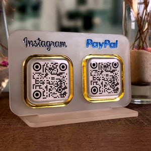 May include: A white acrylic stand with two gold-rimmed square QR code stickers. The left sticker has "Instagram" printed above it and the right sticker has "PayPal" printed above it.