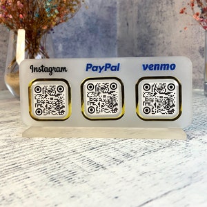 May include: White acrylic sign with three gold-rimmed QR codes. The sign has the words "Instagram", "PayPal", and "venmo" printed on it.