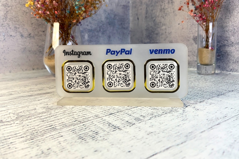 May include: White acrylic sign with three gold-rimmed QR codes. The sign has the words "Instagram", "PayPal", and "venmo" printed on it.