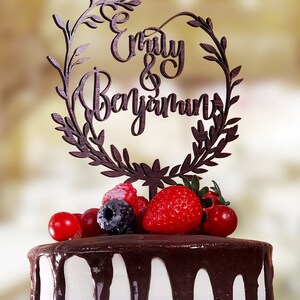 May include: A white cake with chocolate frosting and a wooden cake topper that says "Emily & Benjamin" in a cursive font. The cake topper is decorated with a wreath of leaves. The cake is decorated with fresh berries.