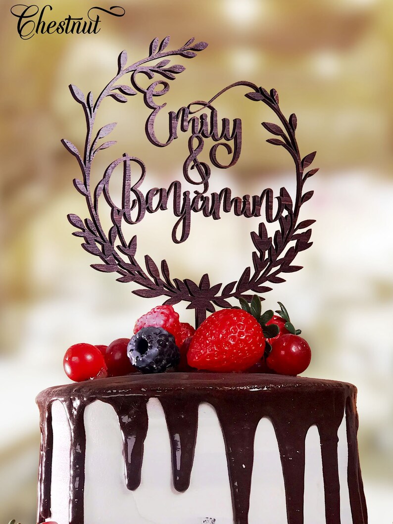 May include: A white cake with chocolate frosting and a wooden cake topper that says "Emily & Benjamin" in a cursive font. The cake topper is decorated with a wreath of leaves. The cake is decorated with fresh berries.