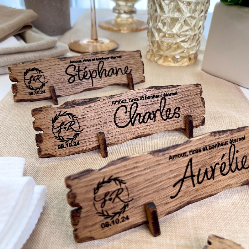 May include: Three rustic wooden place cards with a decorative wreath and initials "S & R" and the date "08.10.24" engraved on each. The place cards are labeled "St&eacute;phane", "Charles", and "Aur&eacute;lie".
