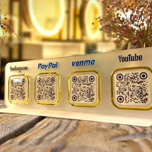 May include: A white stand with four square QR codes, each with a different logo: Instagram, PayPal, Venmo, and YouTube. The stand is on a wooden surface.