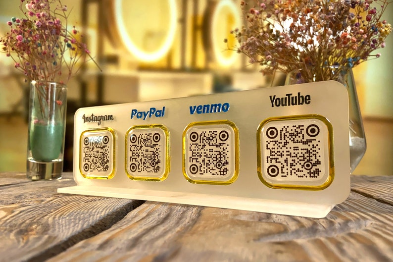 May include: A white stand with four square QR codes, each with a different logo: Instagram, PayPal, Venmo, and YouTube. The stand is on a wooden surface.