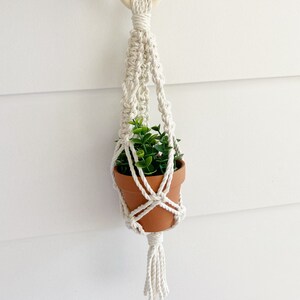 Peut inclure: Un porte-plante en macramé blanc avec un anneau en bois en haut et un pompon en bas. Un petit pot en terre cuite avec une plante verte est suspendu au macramé.