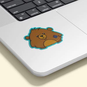 May include: A brown bear sticker with a light blue outline. The bear has a tan belly and a small blue patch on its side. The sticker is on a white laptop.