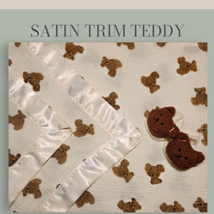 May include: A white muslin baby blanket with a satin trim and a brown teddy bear print. The blanket is folded in half and has a small brown teddy bear attached to it. The text "LITTLE MONSTERS CHICAGO Teddy Bear Collection" is printed on the blanket.