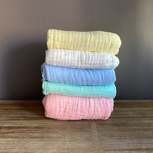 May include: A stack of five soft, textured baby blankets in pastel colours: yellow, white, light blue, mint green, and pink.