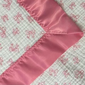 May include: A baby blanket featuring a white, quilted fabric with a pink bow pattern. The blanket is trimmed with a smooth, satin-like pink border. The design is soft and delicate, suitable for a nursery.