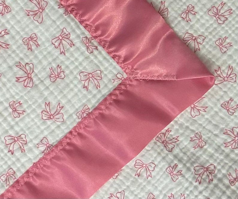 May include: A baby blanket featuring a white, quilted fabric with a pink bow pattern. The blanket is trimmed with a smooth, satin-like pink border. The design is soft and delicate, suitable for a nursery.