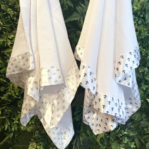 Op de afbeelding: Twee witte babydekens van wafelstof met satijnen rand. De ene deken heeft een patroon van grijze walvissen, de andere heeft marineblauwe ankers. De dekens worden getoond tegen een achtergrond van groen gebladerte.