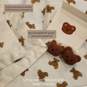 May include: A white muslin baby blanket with a brown teddy bear print. The blanket has a satin border and rounded corners. The text "Double stitched satin for durability ADHD and sensory touch approved" and "Special rounded fold "pocket" corners for sensory feels" is printed on the blanket. The text "LITTLE MONSTERS CHICAGO Teddy Collection" is printed at the bottom of the blanket.