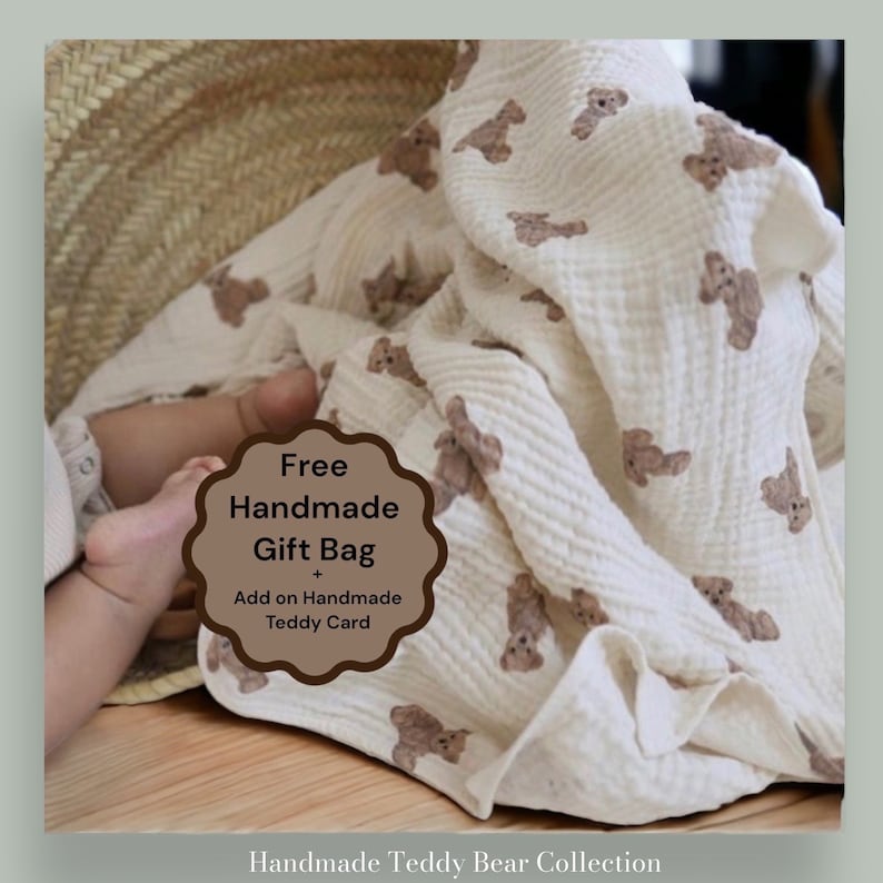 May include: A white muslin baby blanket with a brown teddy bear print. The blanket is folded and draped over a wicker basket. A baby's foot is visible in the lower left corner of the image. The text "Free Handmade Gift Bag + Add on Handmade Teddy Card" is on a brown circle in the center of the image.
