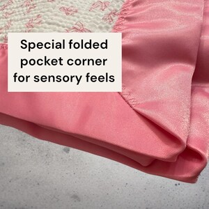 May include: Close-up of a pink fabric with a special folded pocket corner. The fabric has a satin-like texture and is a soft, light pink color. The text on the image reads: "Special folded pocket corner for sensory feels."