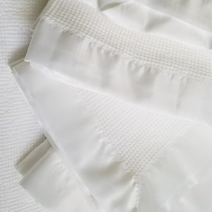 Op de afbeelding: Opgevouwen witte dekens met een wafelweefselpatroon en een gladde satijnen rand. De dekens liggen op een witte, getextureerde ondergrond. De afbeelding toont beddengoed.