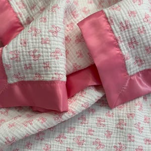 May include: A white baby blanket with a pink bow pattern and a pink satin border. The blanket is made of a soft, textured fabric, perfect for a nursery or as a baby shower gift.