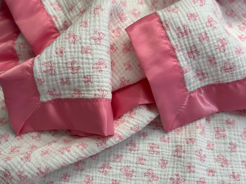 May include: A white baby blanket with a pink bow pattern and a pink satin border. The blanket is made of a soft, textured fabric, perfect for a nursery or as a baby shower gift.