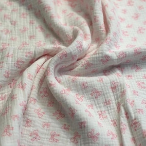 May include: White muslin fabric with a pattern of small pink bows. The fabric is soft and textured, with visible folds and wrinkles. The bows are evenly spaced across the surface, creating a delicate and feminine design.