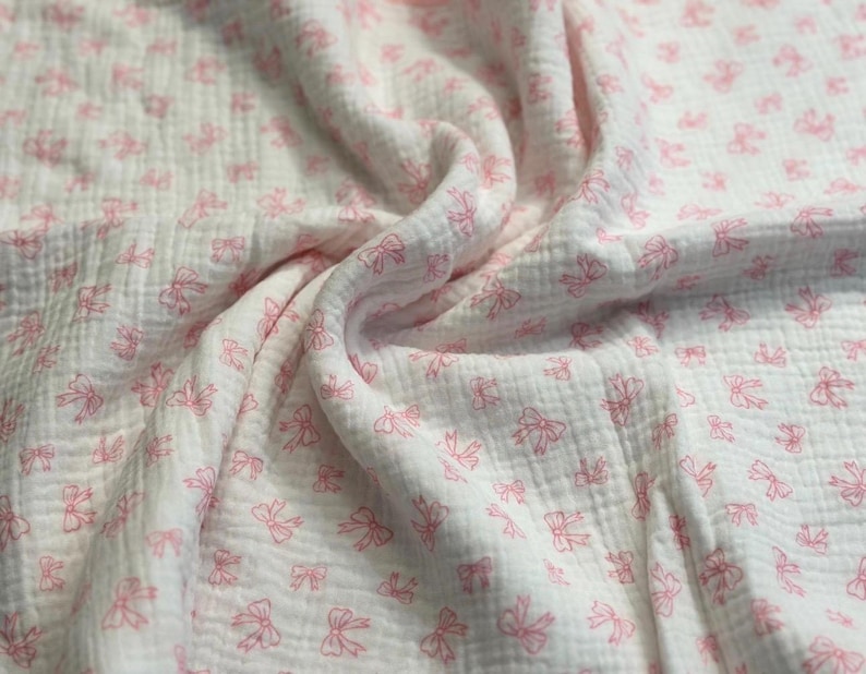 May include: White muslin fabric with a pattern of small pink bows. The fabric is soft and textured, with visible folds and wrinkles. The bows are evenly spaced across the surface, creating a delicate and feminine design.