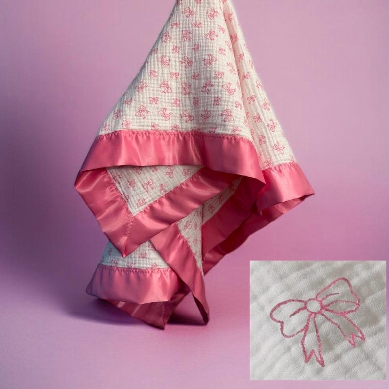 May include: A white and pink baby blanket with a pink satin border. The blanket features a repeating pattern of pink bows and is hanging against a purple background. The satin border adds a touch of elegance to the soft, textured blanket.