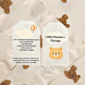 May include: Two white tags with rounded corners and a hole at the top. The tag on the left has a drawing of a hot air balloon and a cloud. The text on the tag reads: "The handmade teddy bear blanket is pre-washed with hypoallergenic fragrance-free detergent and is ready for immediate snuggles. Please leave a 5 star review if you love it and follow us on Instagram @little_monsters_chicago". The tag on the right has a drawing of a brown teddy bear with a white face and a white cloud. The text on the tag reads: "Little Monsters Chicago".