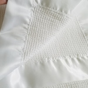Op de afbeelding: Close-up van een witte babydeken met een getextureerd midden en een satijnen rand. De deken is gemaakt van zachte, pluche materialen en heeft gedetailleerde stiksels.