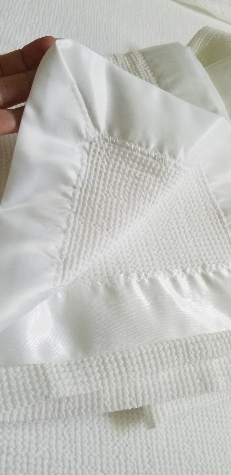 Op de afbeelding: Close-up van een witte babydeken met een getextureerd midden en een satijnen rand. De deken is gemaakt van zachte, pluche materialen en heeft gedetailleerde stiksels.